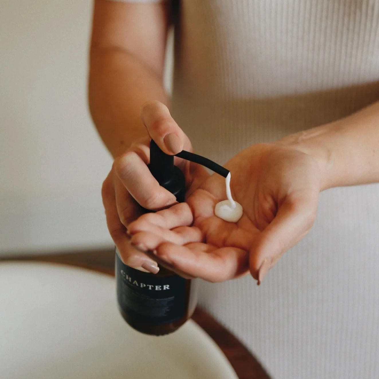A person in a fitted, light-coloured dress is dispensing a small amount of cream from a dark bottle into their palm. The setting appears to be a bathroom, with a soft-focus background. The focus is on the hands and the product.