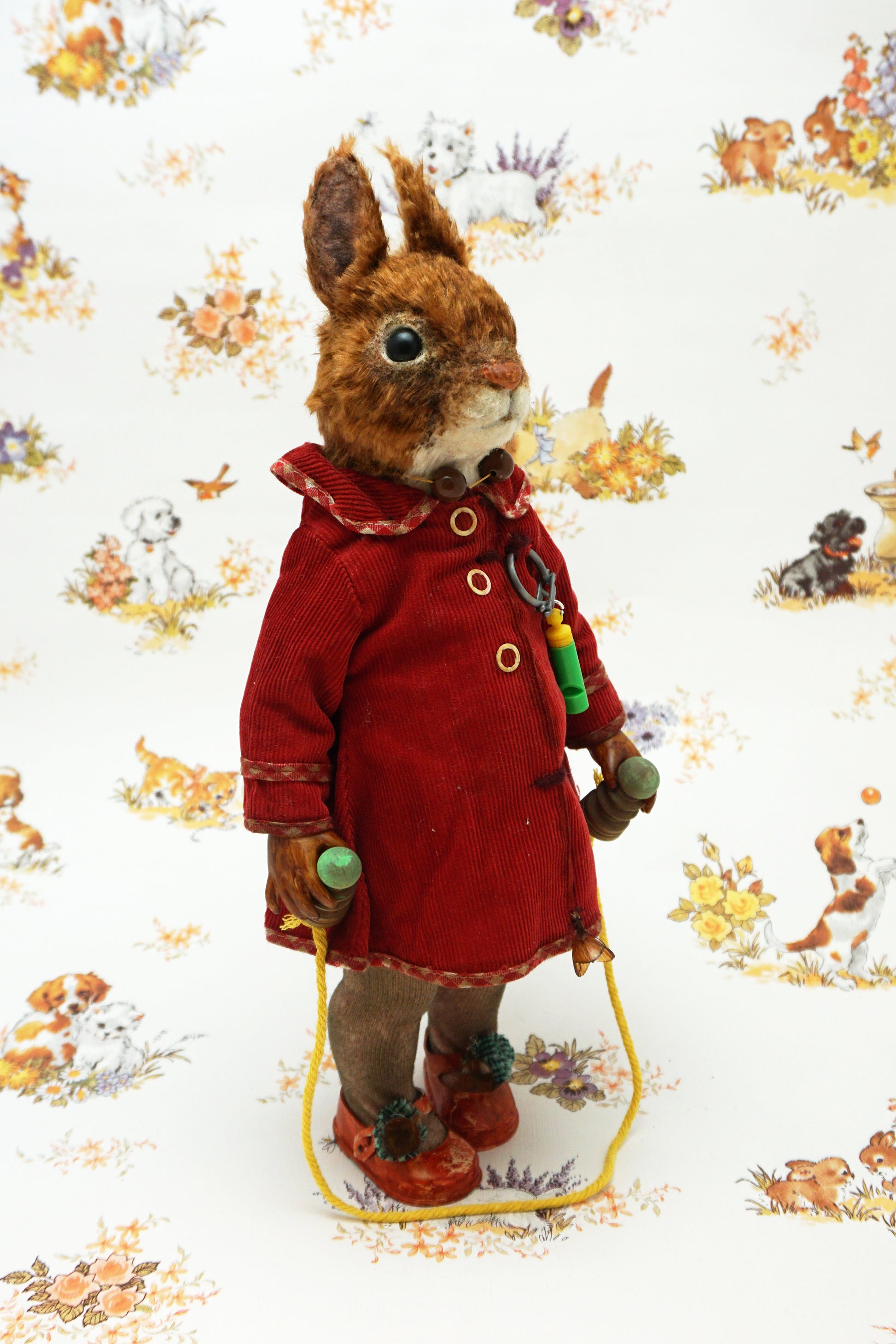 A plush squirrel stands upright, wearing a red coat and holding a skipping rope. The background features a whimsical pattern with small animals and floral designs. The squirrel has a whistle keychain attached to its coat.