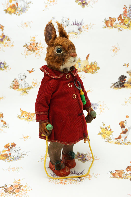 A plush squirrel stands upright, wearing a red coat and holding a skipping rope. The background features a whimsical pattern with small animals and floral designs. The squirrel has a whistle keychain attached to its coat.