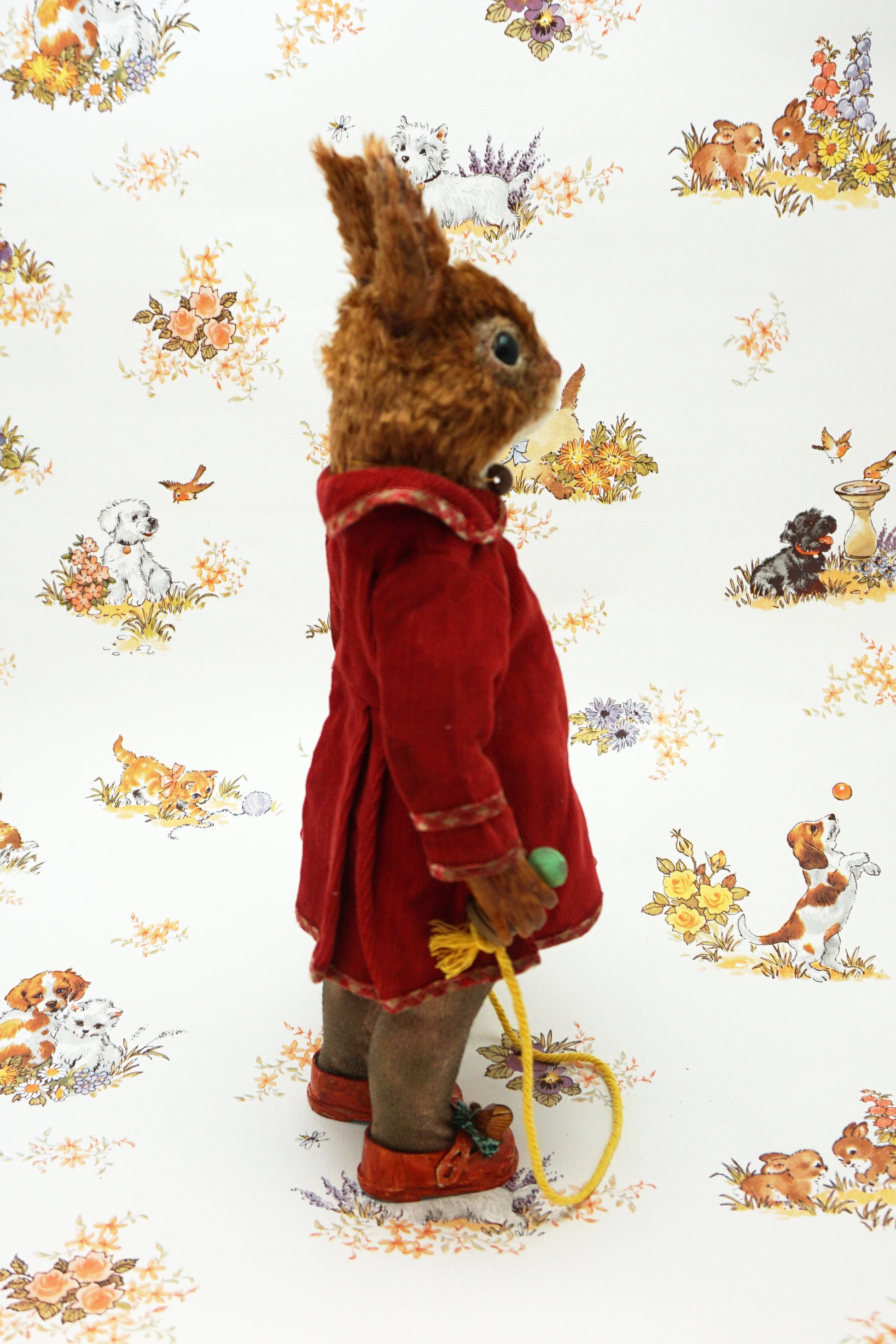 A plush squirrel stands upright, wearing a red coat and holding a skipping rope. The background features a whimsical pattern with small animals and floral designs. The squirrel has a whistle keychain attached to its coat.