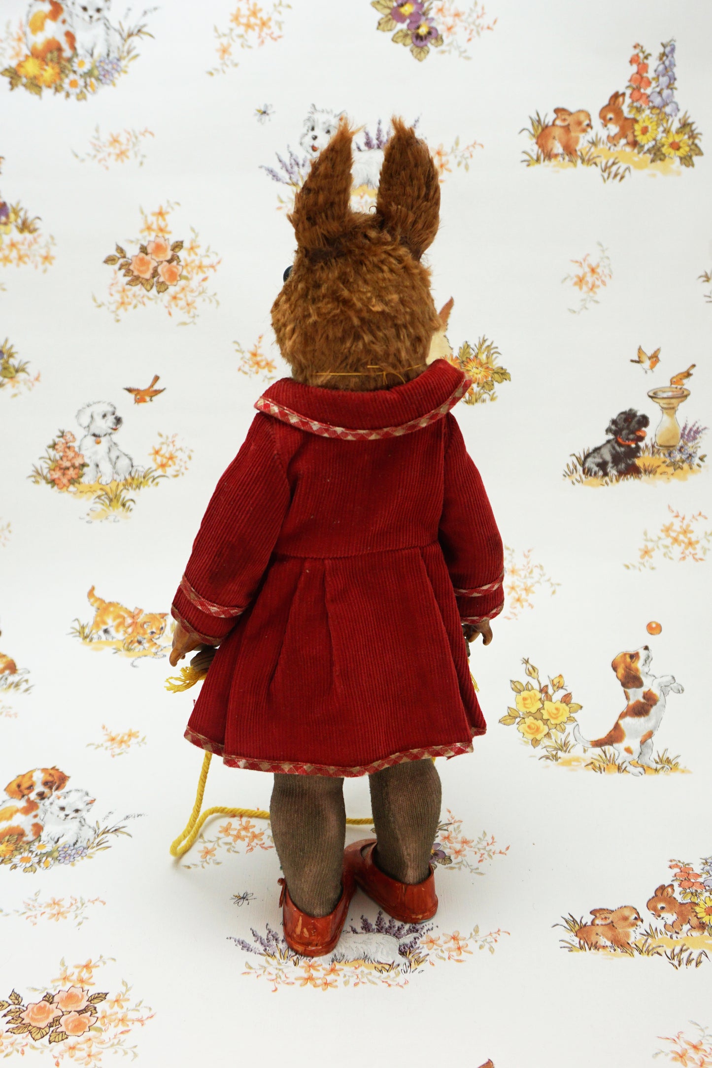 A plush squirrel stands upright, wearing a red coat and holding a skipping rope. The background features a whimsical pattern with small animals and floral designs. The squirrel has a whistle keychain attached to its coat.