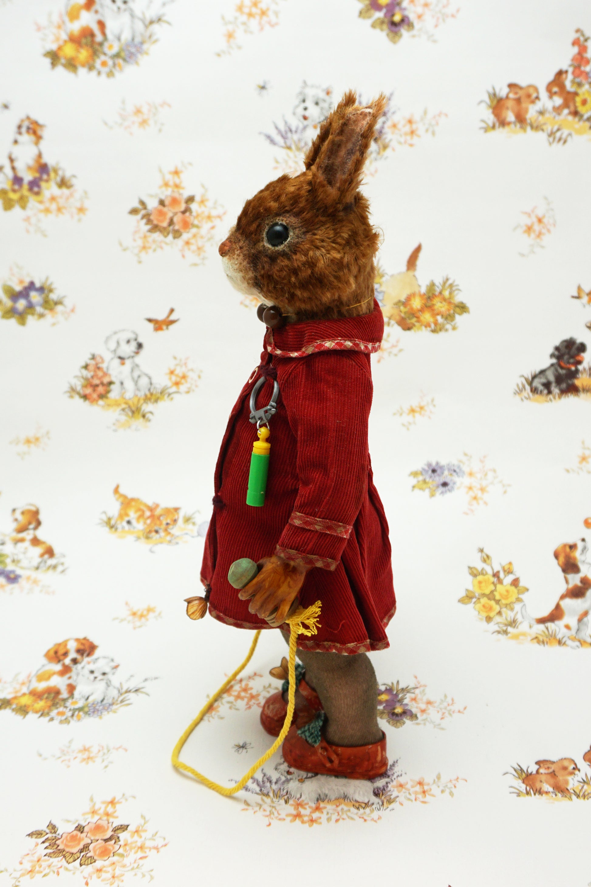 A plush squirrel stands upright, wearing a red coat and holding a skipping rope. The background features a whimsical pattern with small animals and floral designs. The squirrel has a whistle keychain attached to its coat.