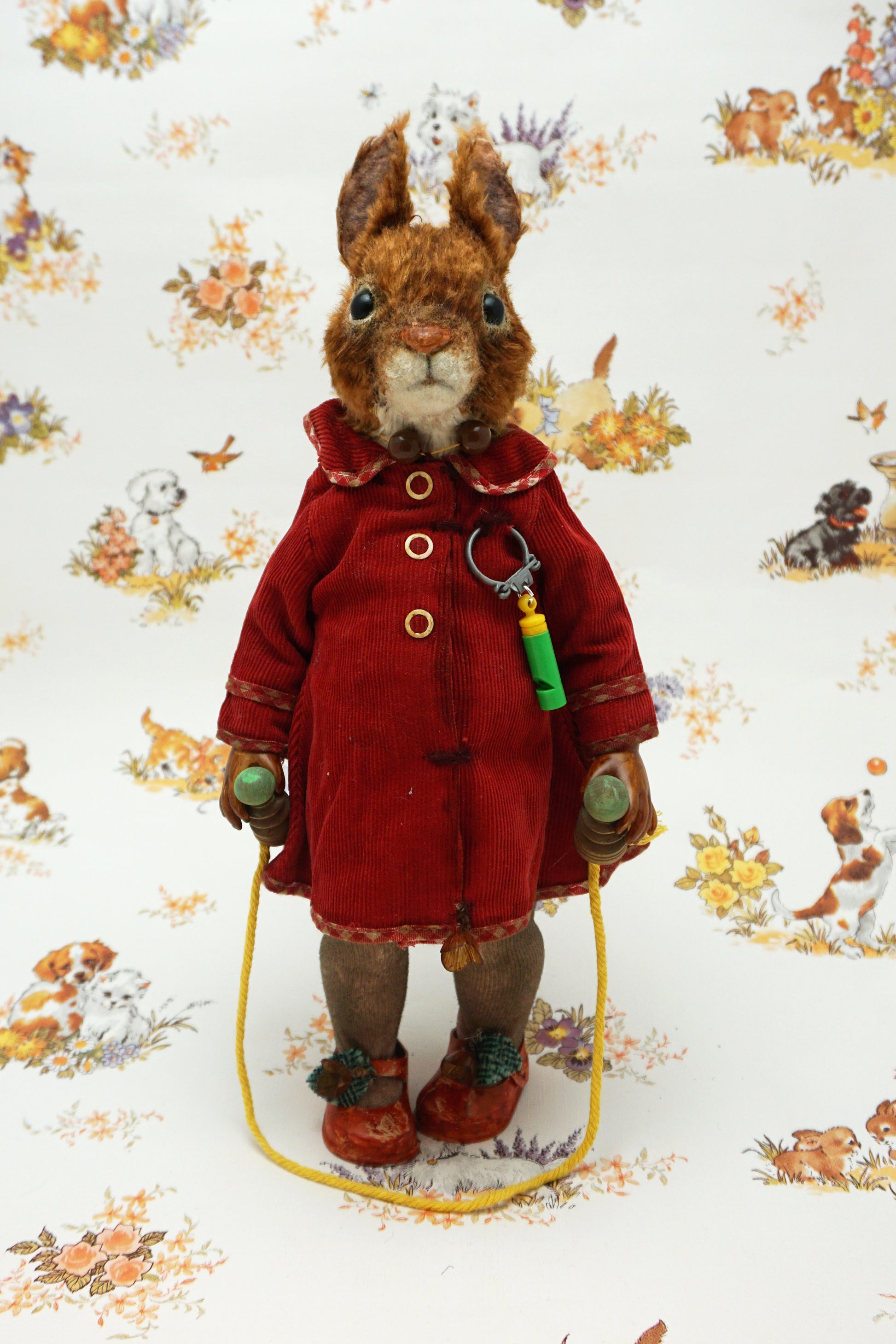 A plush squirrel stands upright, wearing a red coat and holding a skipping rope. The background features a whimsical pattern with small animals and floral designs. The squirrel has a whistle keychain attached to its coat.