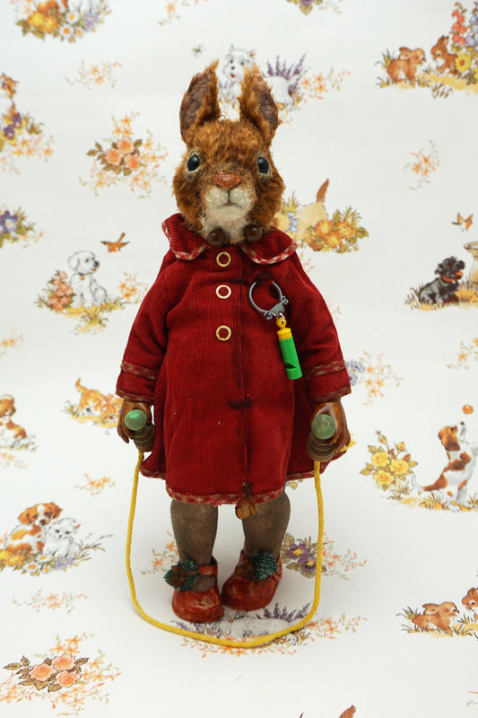 A plush squirrel stands upright, wearing a red coat and holding a skipping rope. The background features a whimsical pattern with small animals and floral designs. The squirrel has a whistle keychain attached to its coat.