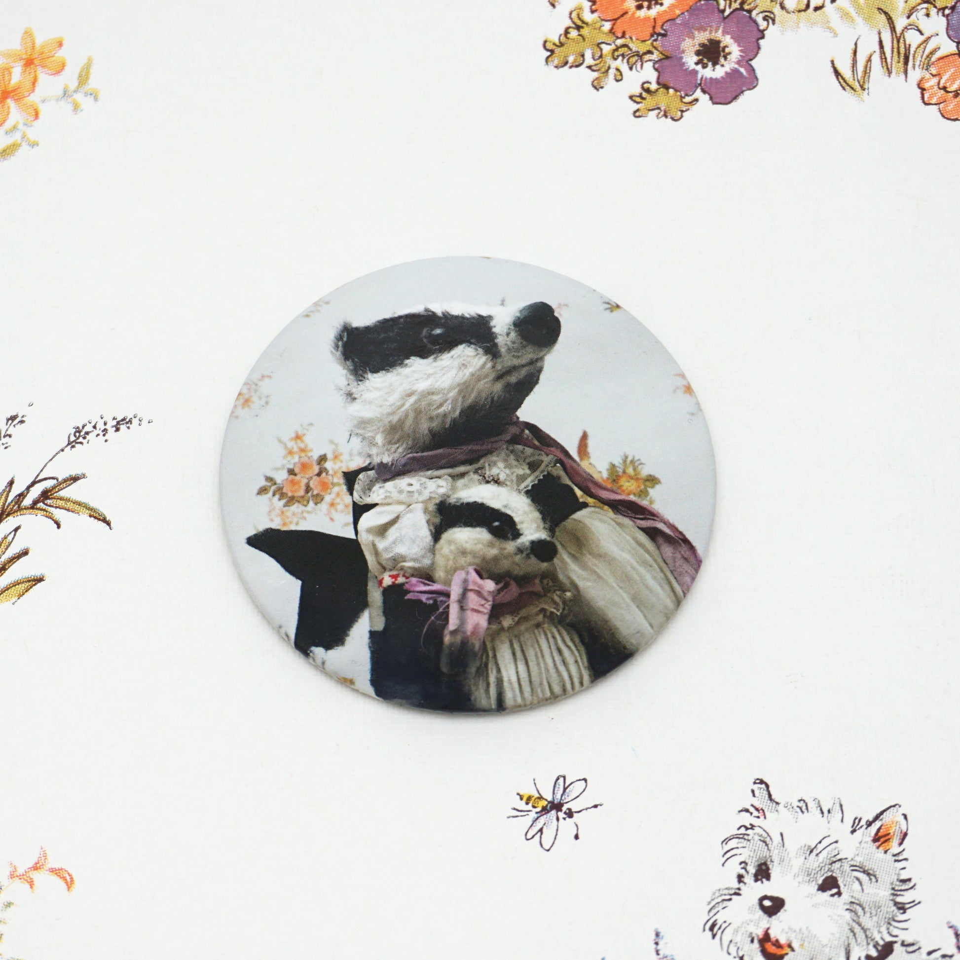 A round pocket mirror features two badgers dressed in vintage clothing, with floral patterns in the background. The larger badger wears a scarf, while the smaller one is wrapped in a pink ribbon. The badge is placed on a floral background.