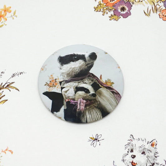A round pocket mirror features two badgers dressed in vintage clothing, with floral patterns in the background. The larger badger wears a scarf, while the smaller one is wrapped in a pink ribbon. The badge is placed on a floral background.