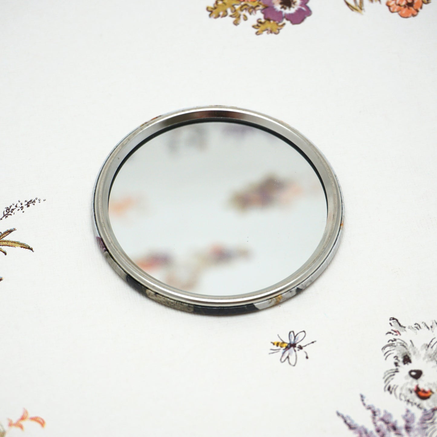 A round, silver-framed mirror lies on a white surface adorned with colourful floral patterns and a small dog illustration. The mirror reflects a blurred portion of the floral design.