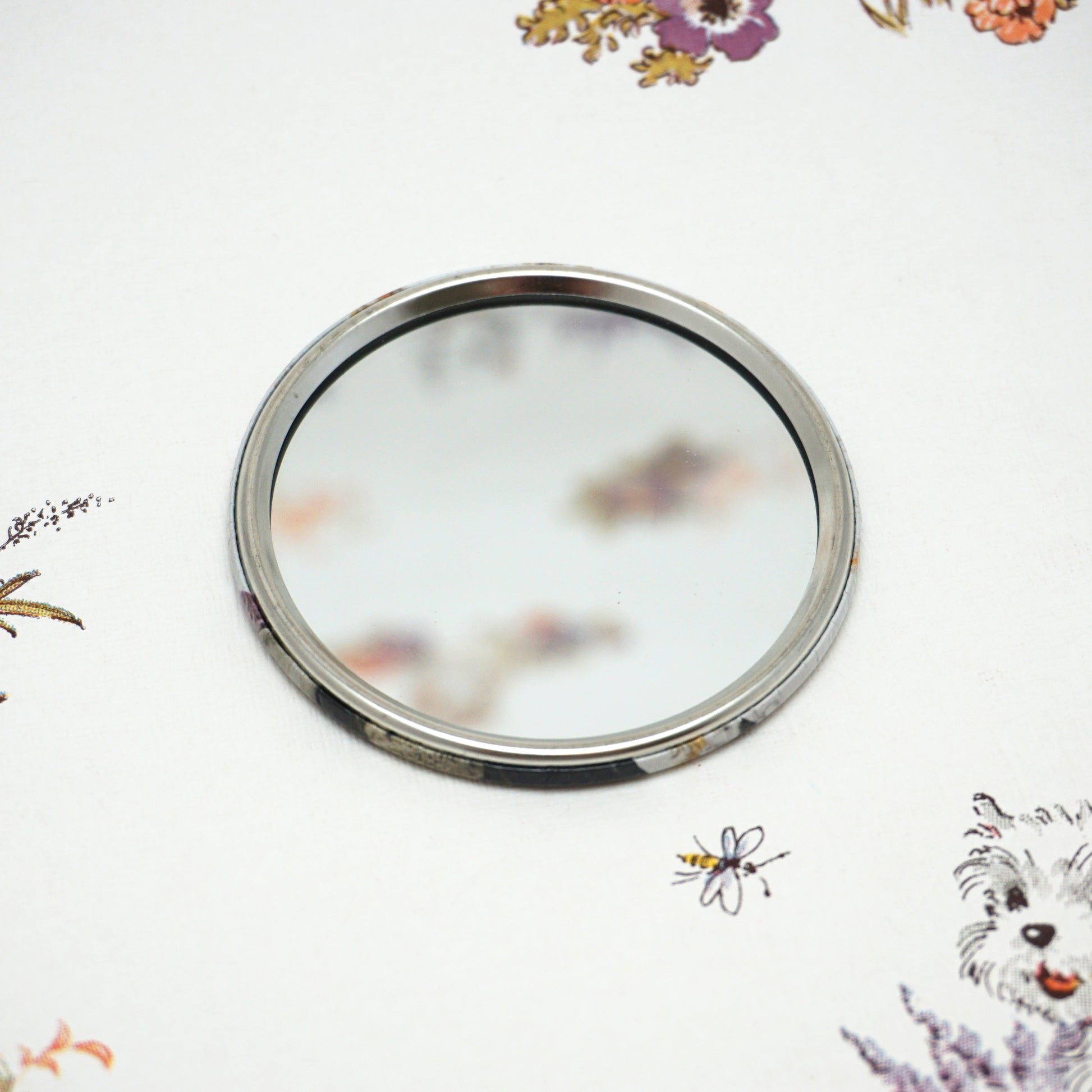 A round, silver-framed mirror lies on a white surface adorned with colourful floral patterns and a small dog illustration. The mirror reflects a blurred portion of the floral design.