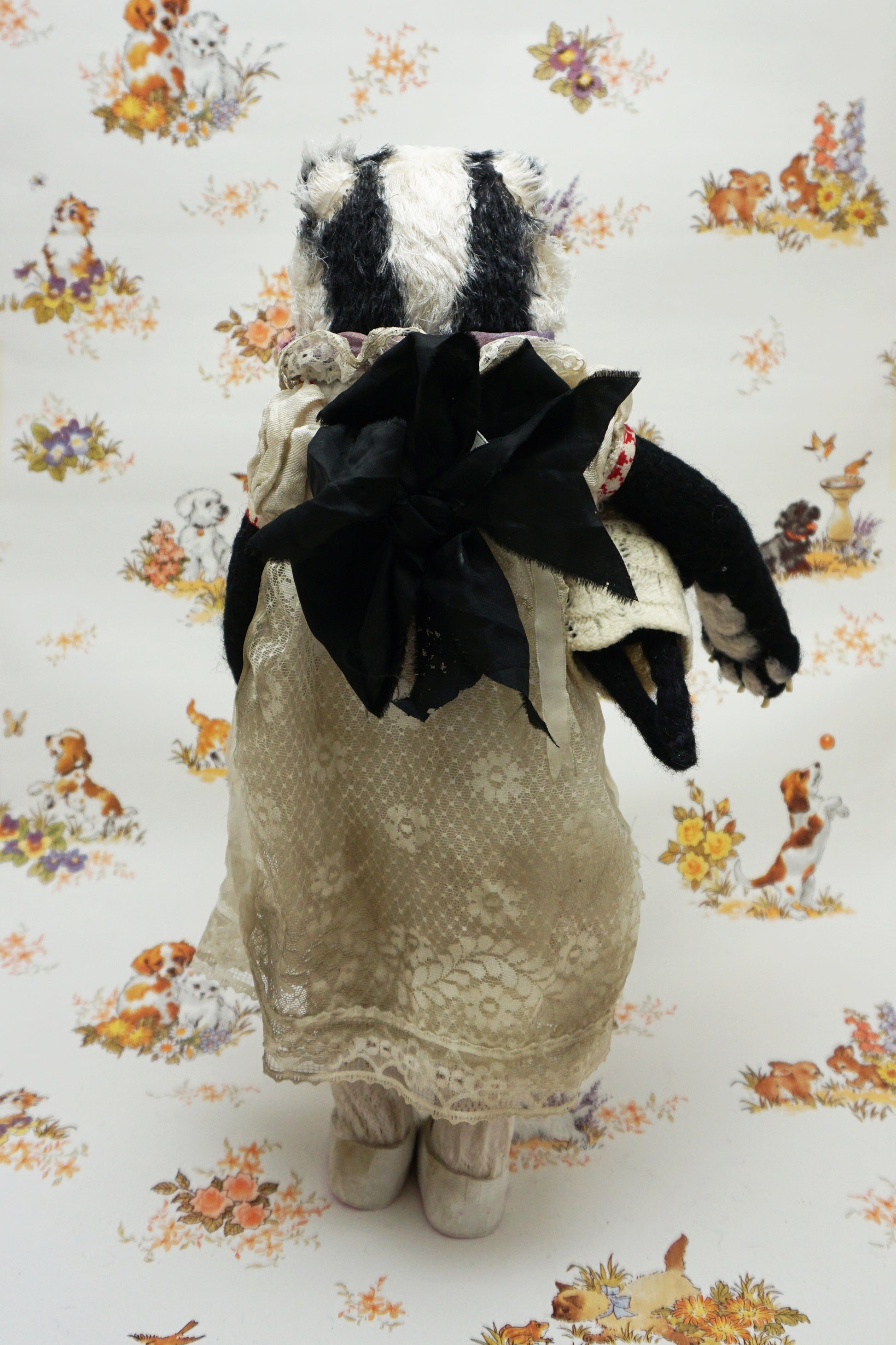 A plush badger toy dressed in a vintage-style lace dress and purple scarf stands holding a smaller badger toy in similar attire. The background features a whimsical pattern with animals and flowers.