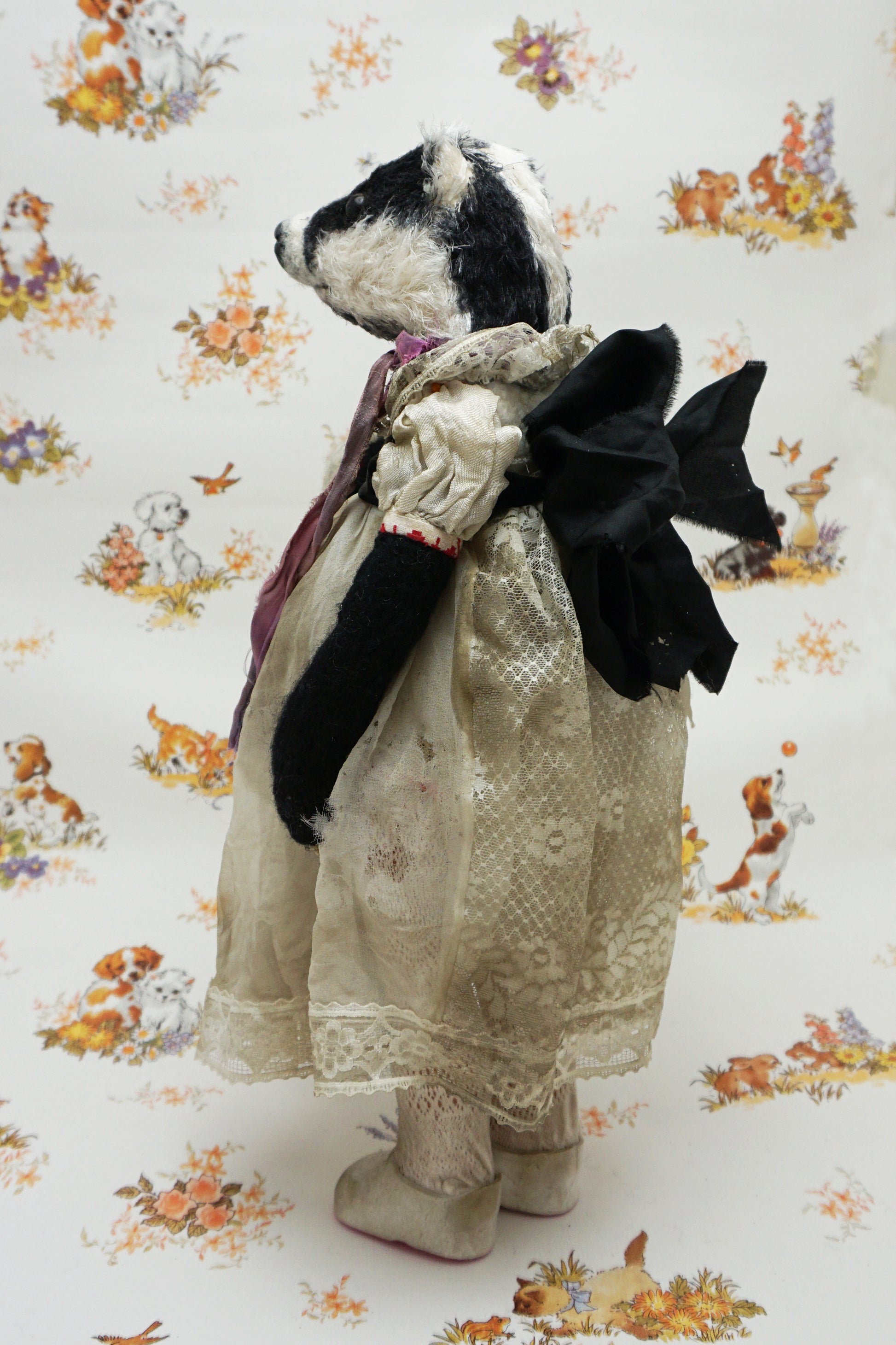 A plush badger toy dressed in a vintage-style lace dress and purple scarf stands holding a smaller badger toy in similar attire. The background features a whimsical pattern with animals and flowers.