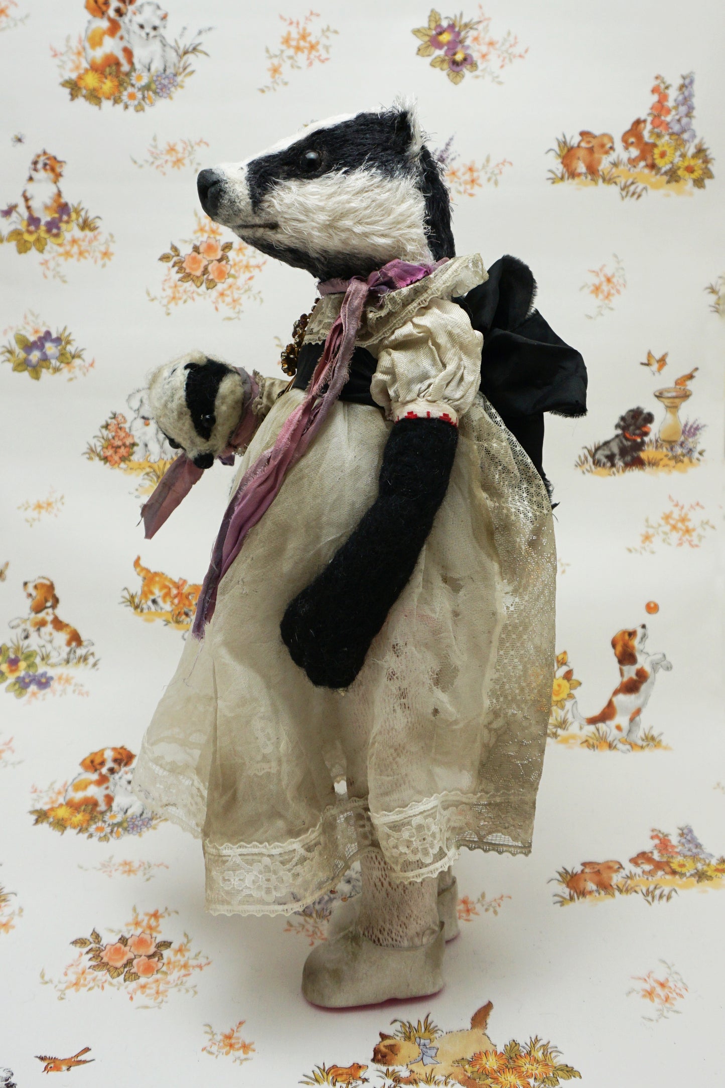 A plush badger toy dressed in a vintage-style lace dress and purple scarf stands holding a smaller badger toy in similar attire. The background features a whimsical pattern with animals and flowers.