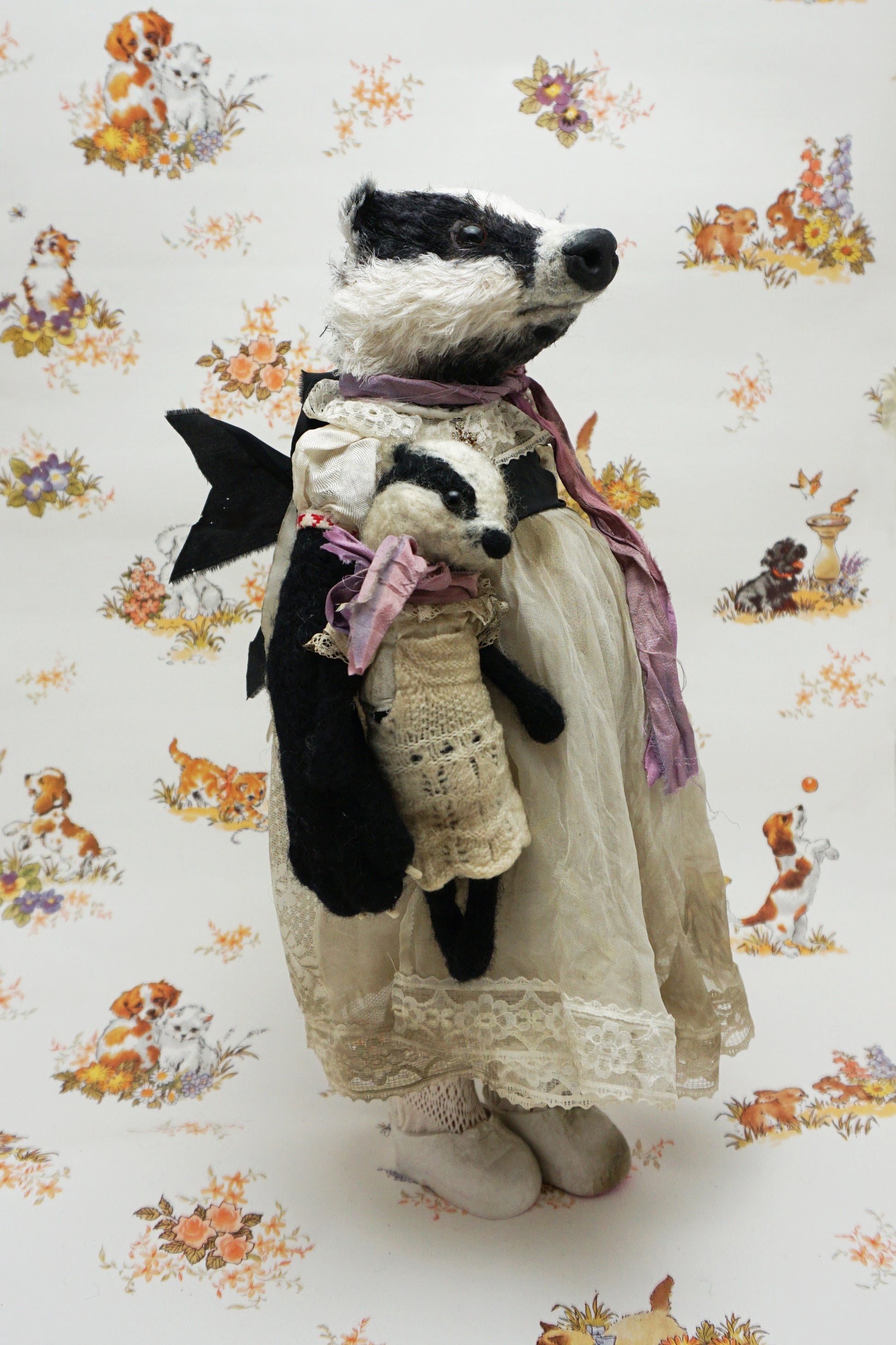 A plush badger toy dressed in a vintage-style lace dress and purple scarf stands holding a smaller badger toy in similar attire. The background features a whimsical pattern with animals and flowers.