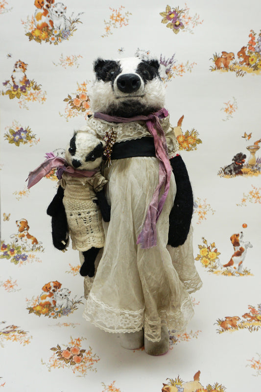 A plush badger toy dressed in a vintage-style lace dress and purple scarf stands holding a smaller badger toy in similar attire. The background features a whimsical pattern with animals and flowers.