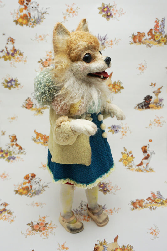 A whimsical felted dog sculpture stands upright, wearing a blue dress with a beige coat. It has a fluffy mane and a cheerful expression. The background features a playful pattern of animals and flowers.