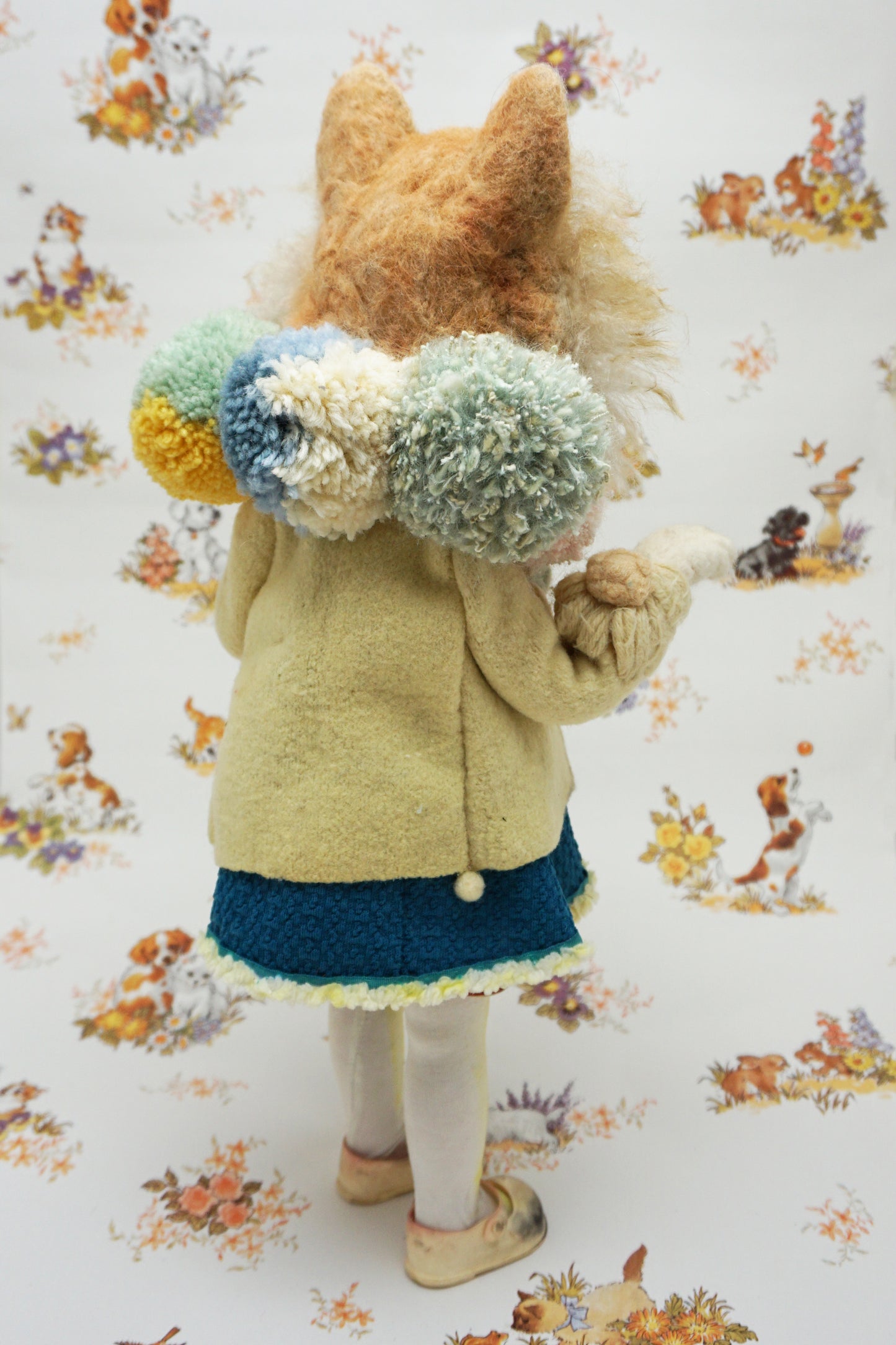 A whimsical felted dog sculpture stands upright, wearing a blue dress with a beige coat. It has a fluffy mane and a cheerful expression. The background features a playful pattern of animals and flowers.
