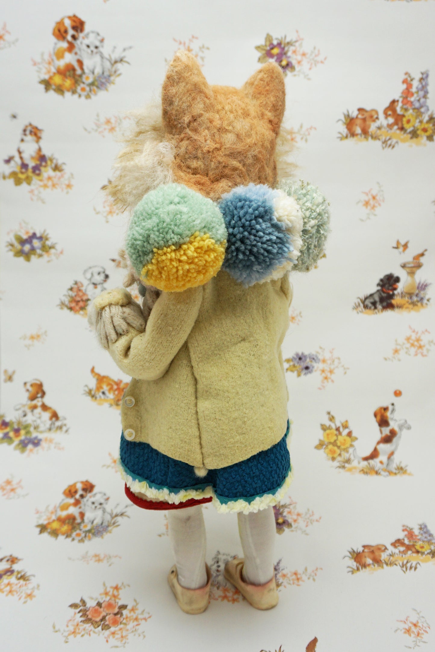 A whimsical felted dog sculpture stands upright, wearing a blue dress with a beige coat. It has a fluffy mane and a cheerful expression. The background features a playful pattern of animals and flowers.