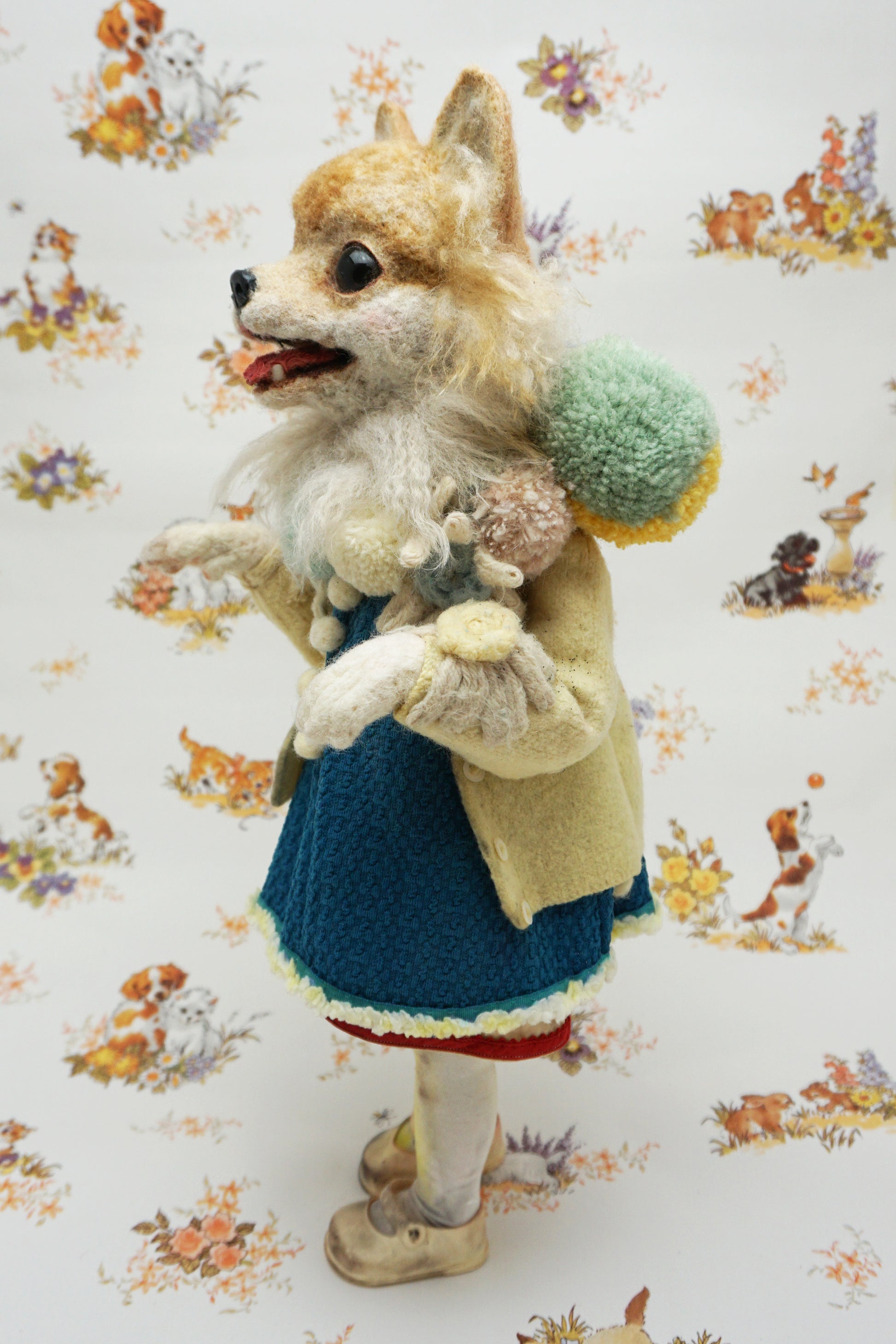 A whimsical felted dog sculpture stands upright, wearing a blue dress with a beige coat. It has a fluffy mane and a cheerful expression. The background features a playful pattern of animals and flowers.