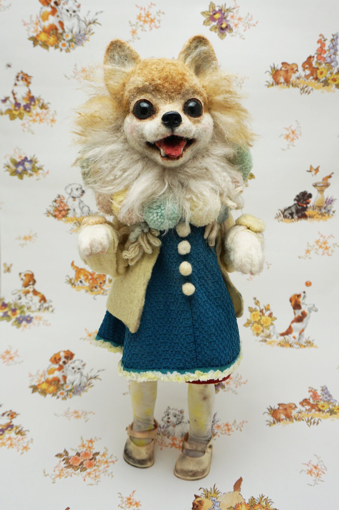 A whimsical felted dog sculpture stands upright, wearing a blue dress with a beige coat. It has a fluffy mane and a cheerful expression. The background features a playful pattern of animals and flowers.