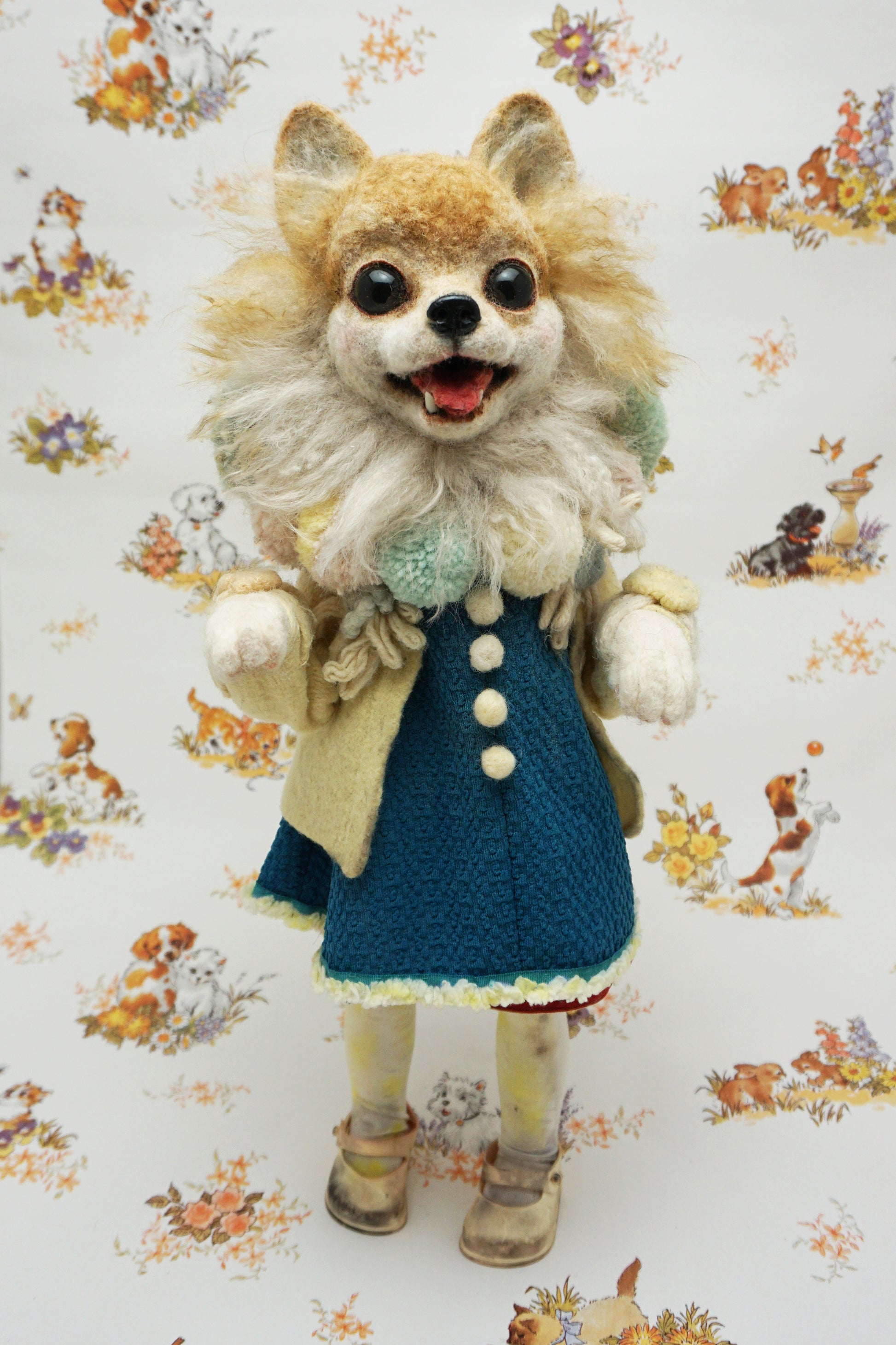 A whimsical felted dog sculpture stands upright, wearing a blue dress with a beige coat. It has a fluffy mane and a cheerful expression. The background features a playful pattern of animals and flowers.