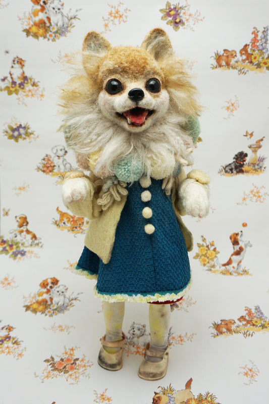 A whimsical felted dog sculpture stands upright, wearing a blue dress with a beige coat. It has a fluffy mane and a cheerful expression. The background features a playful pattern of animals and flowers.