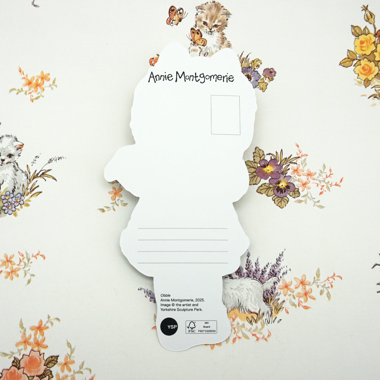 A decorative card featuring a cut-out silhouette of a figure with floral and animal illustrations. The background is adorned with vintage-style flowers and kittens. The name "Annie Montgomerie" is printed at the top.