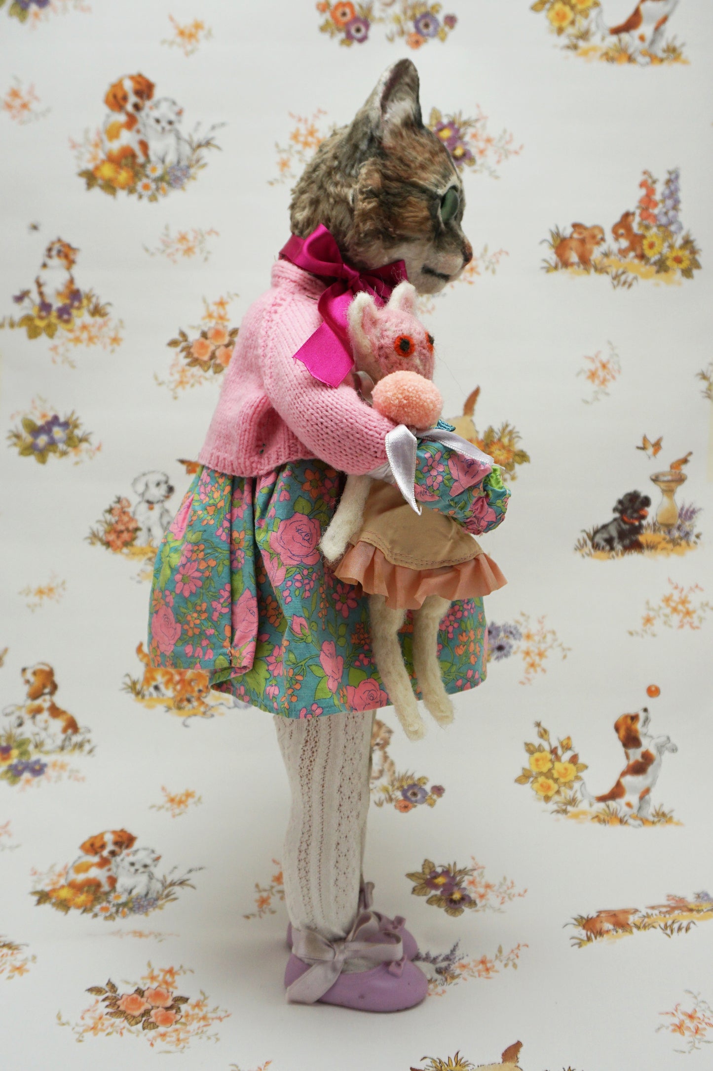 A whimsical cat doll dressed in a floral dress, pink cardigan, and purple shoes holds two smaller plush toys. The background features playful animal illustrations, adding a charming, vintage feel to the scene.