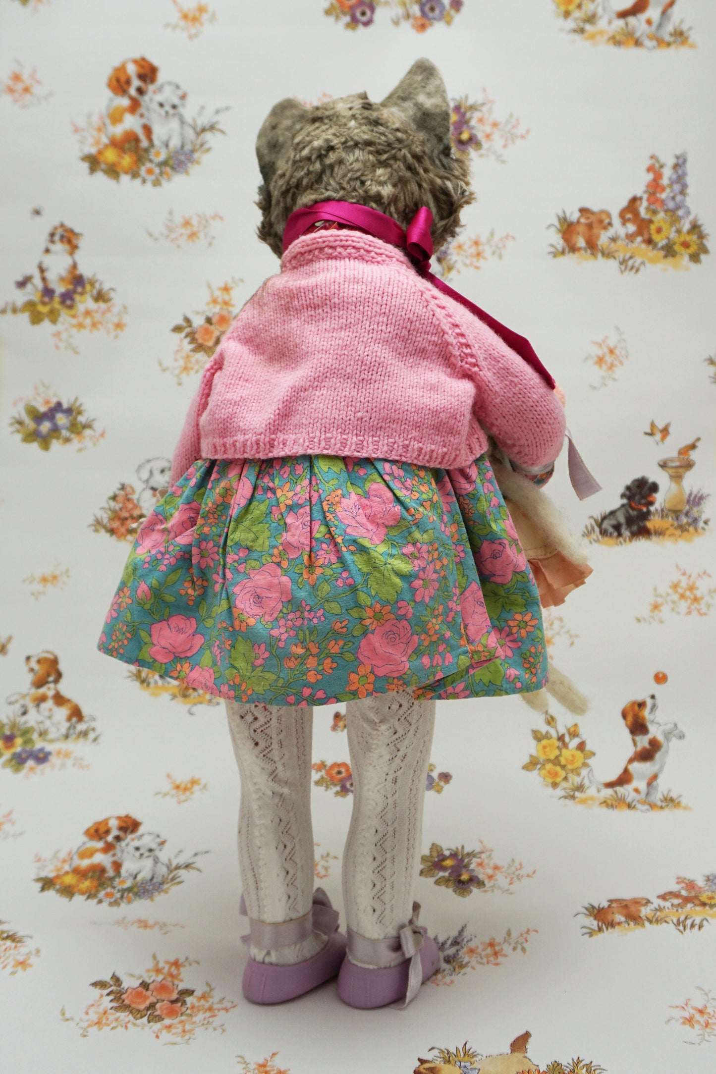 A whimsical cat doll dressed in a floral dress, pink cardigan, and purple shoes holds two smaller plush toys. The background features playful animal illustrations, adding a charming, vintage feel to the scene.