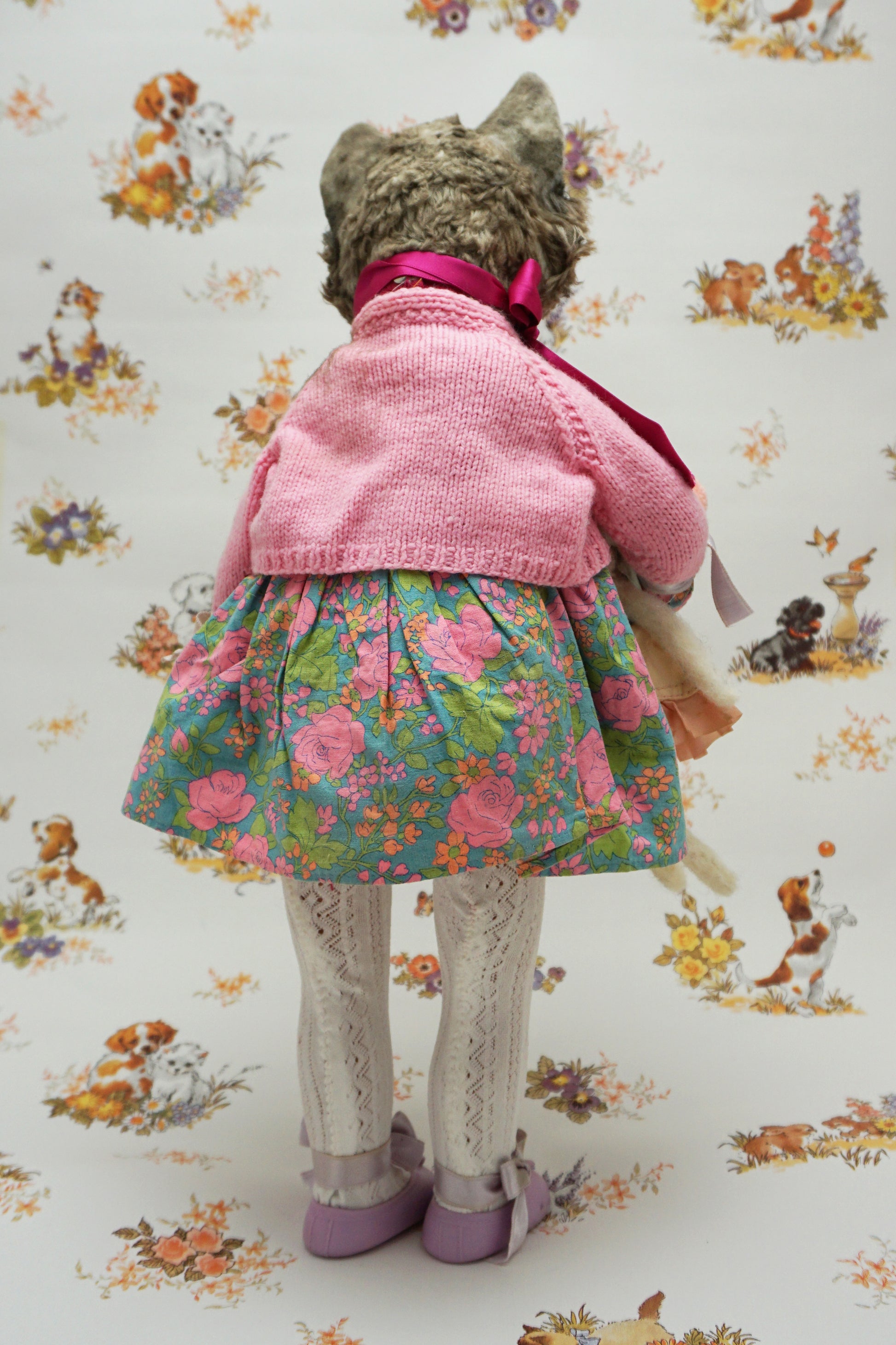 A whimsical cat doll dressed in a floral dress, pink cardigan, and purple shoes holds two smaller plush toys. The background features playful animal illustrations, adding a charming, vintage feel to the scene.