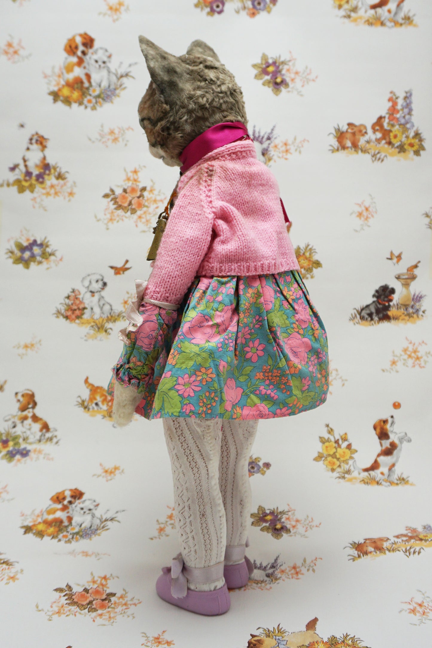 A whimsical cat doll dressed in a floral dress, pink cardigan, and purple shoes holds two smaller plush toys. The background features playful animal illustrations, adding a charming, vintage feel to the scene.