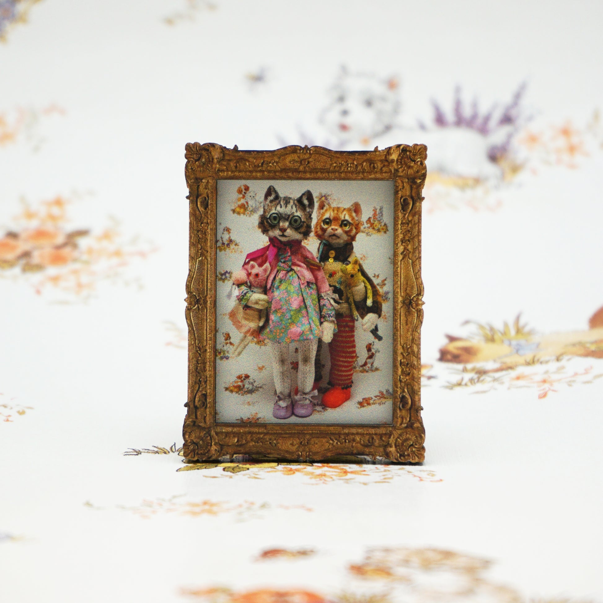 A small ornate frame displays an illustration of two anthropomorphic cats dressed in colourful clothing. One cat holds a pink toy, and the background features a floral pattern. The frame is set against a blurred, similar floral backdrop.