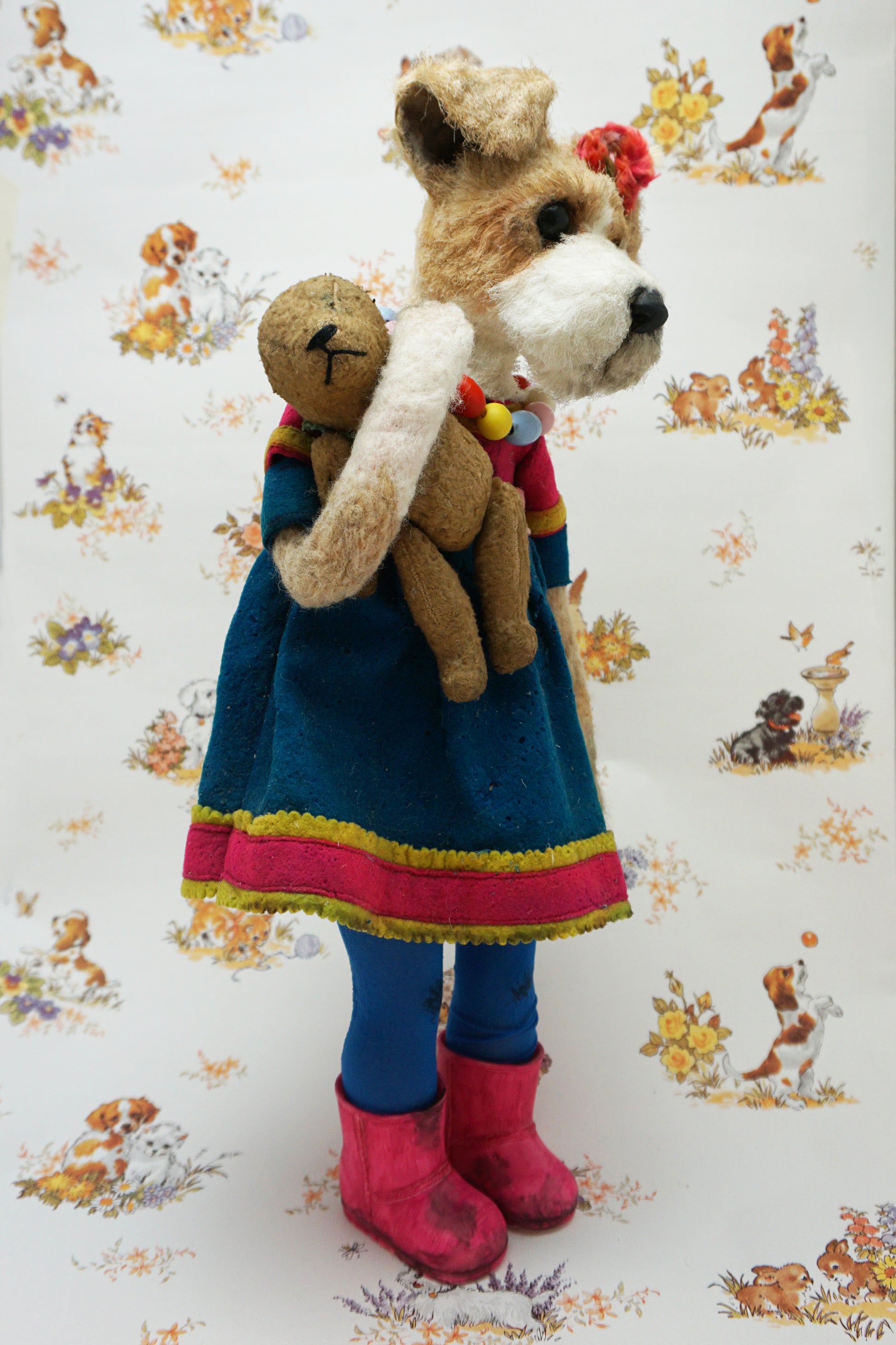 A plush dog dressed in a blue dress with pink trim, colourful bead necklace, and pink boots, holding a smaller plush bear. The background features a pattern of various playful animals and floral designs.
