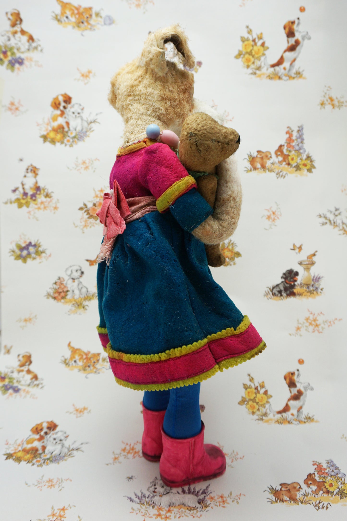 A plush dog dressed in a blue dress with pink trim, colourful bead necklace, and pink boots, holding a smaller plush bear. The background features a pattern of various playful animals and floral designs.