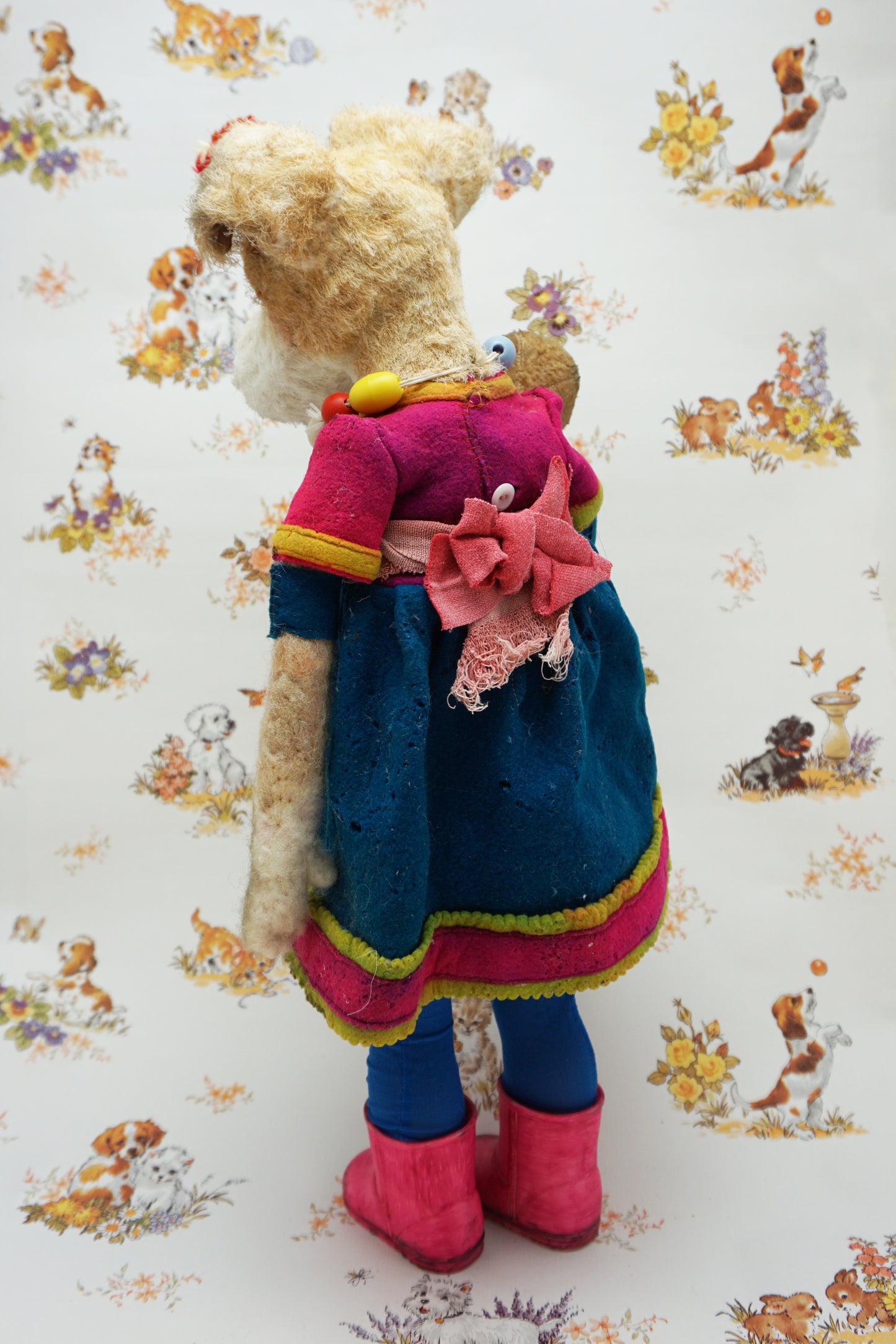 A plush dog dressed in a blue dress with pink trim, colourful bead necklace, and pink boots, holding a smaller plush bear. The background features a pattern of various playful animals and floral designs.