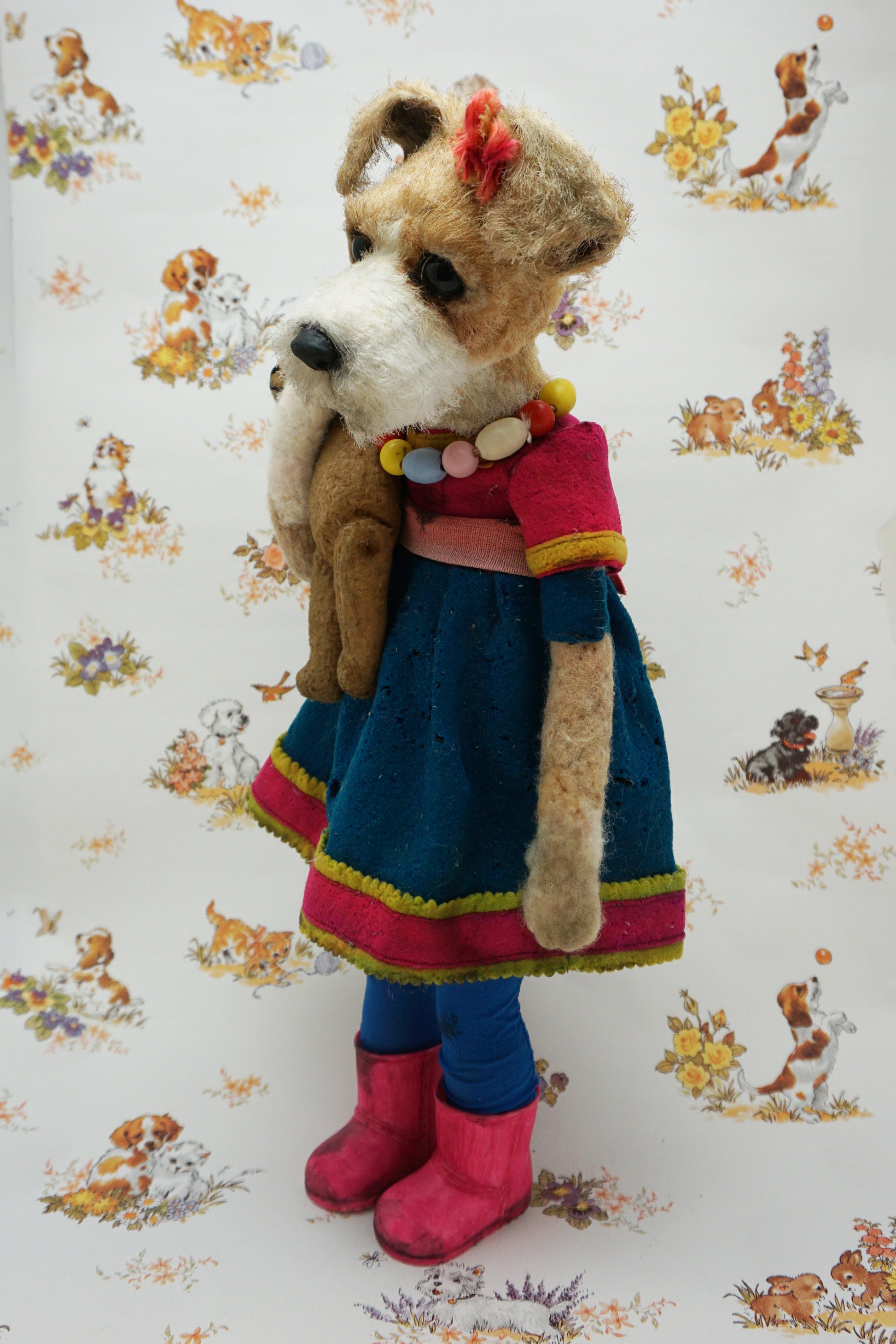 A plush dog dressed in a blue dress with pink trim, colourful bead necklace, and pink boots, holding a smaller plush bear. The background features a pattern of various playful animals and floral designs.