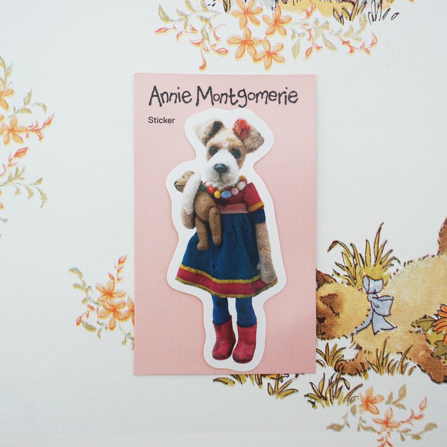 A die-cut sticker featuring a dog character dressed in a blue dress with red trim and red boots, holding a teddy bear, displayed on a pink backing card with the text 'Annie Montgomerie Sticker'. The card is placed on a white background with a floral and illustrated pattern.