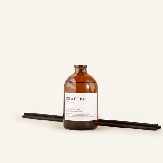 A brown glass bottle labelled "CHAPTER ORGANICS" sits on a light background. Two black incense sticks lie beside the bottle, creating a minimalist and calming aesthetic.