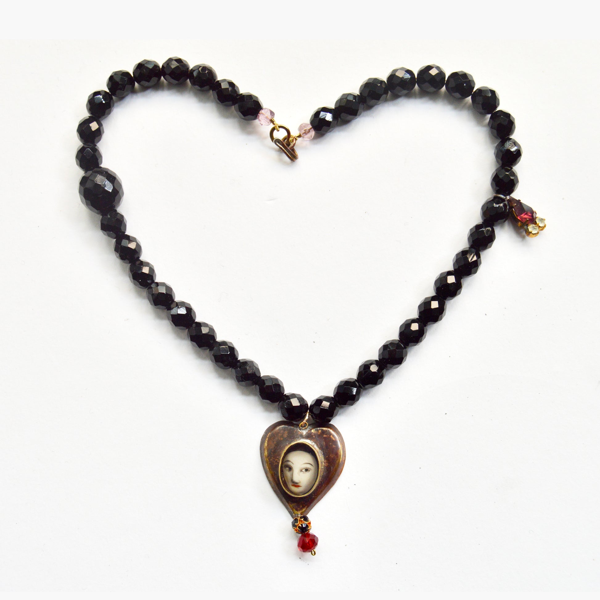A neckpiece with a black beaded chain, a pendant in a heart shape with a ladies face inside, with two bead charms hanging down.