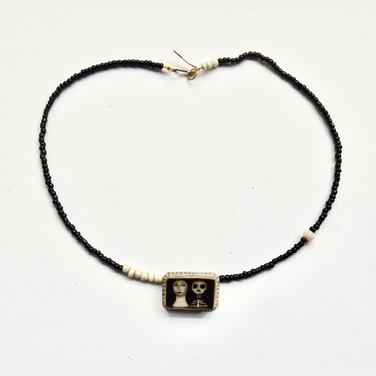 A necklace with a thin beaded chain the beads are black and white. The pendant attached at the front shows an illustration of a women's neck and face next to a skeletons head and ribs. 