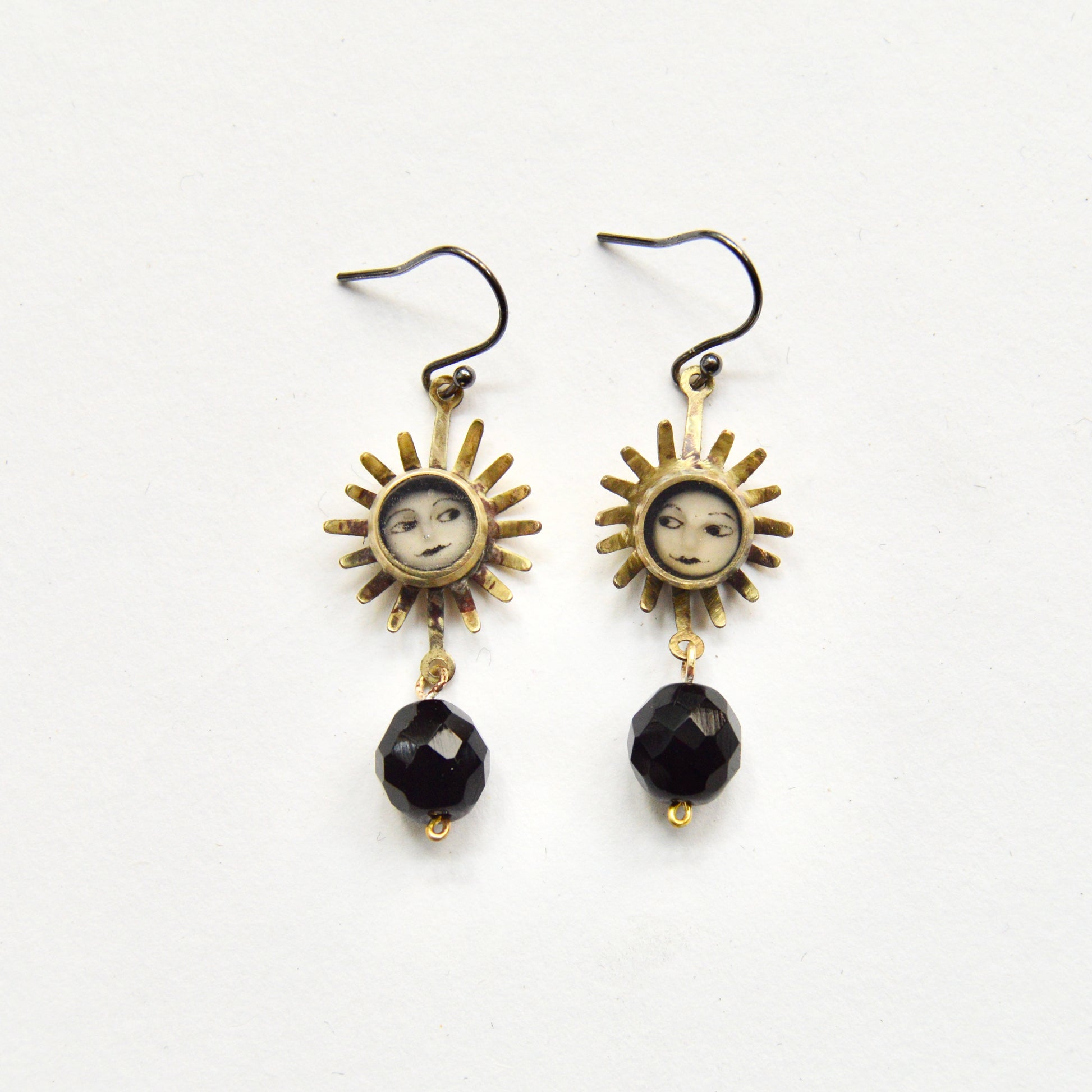 A pair of hanging earrings featuring a sun with a painted face inset on each earring. Under the sun dangles a single black bead. 