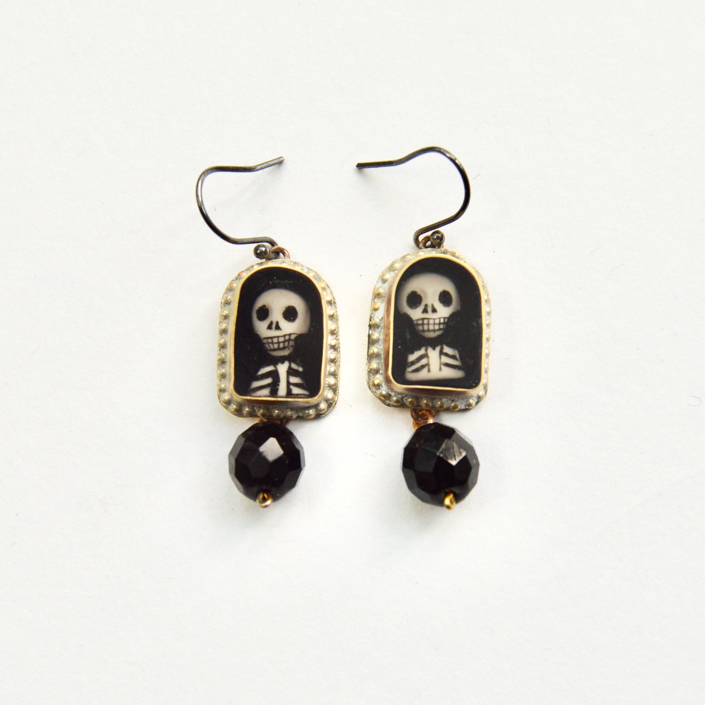 A pair of hanging earrings, each featuring a golden arch with a hand painted insert of a skeletons head and torse. Under each arch hangs a single glass black bead.