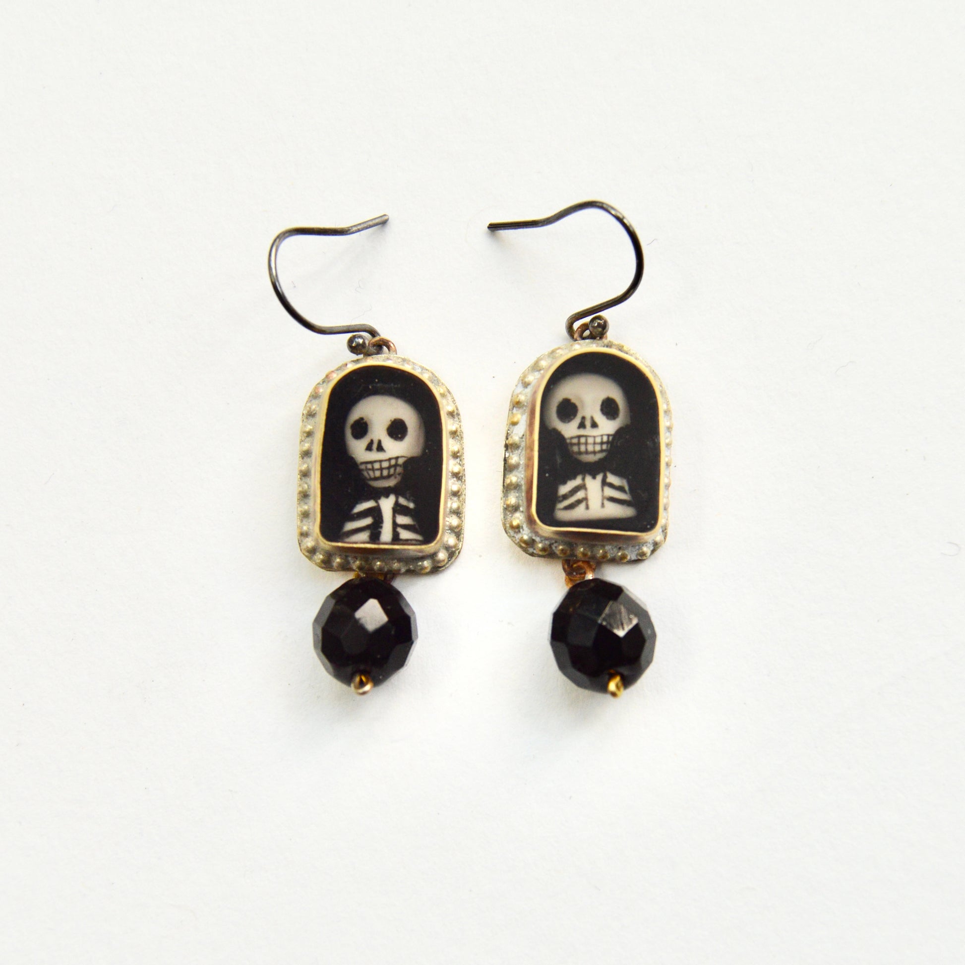 A pair of hanging earrings, each featuring a golden arch with a hand painted insert of a skeletons head and torse. Under each arch hangs a single glass black bead.