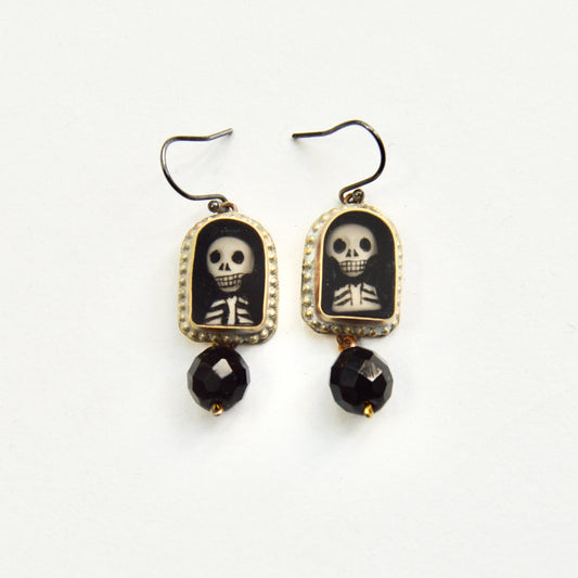 A pair of hanging earrings, each featuring a golden arch with a hand painted insert of a skeletons head and torse. Under each arch hangs a single glass black bead.