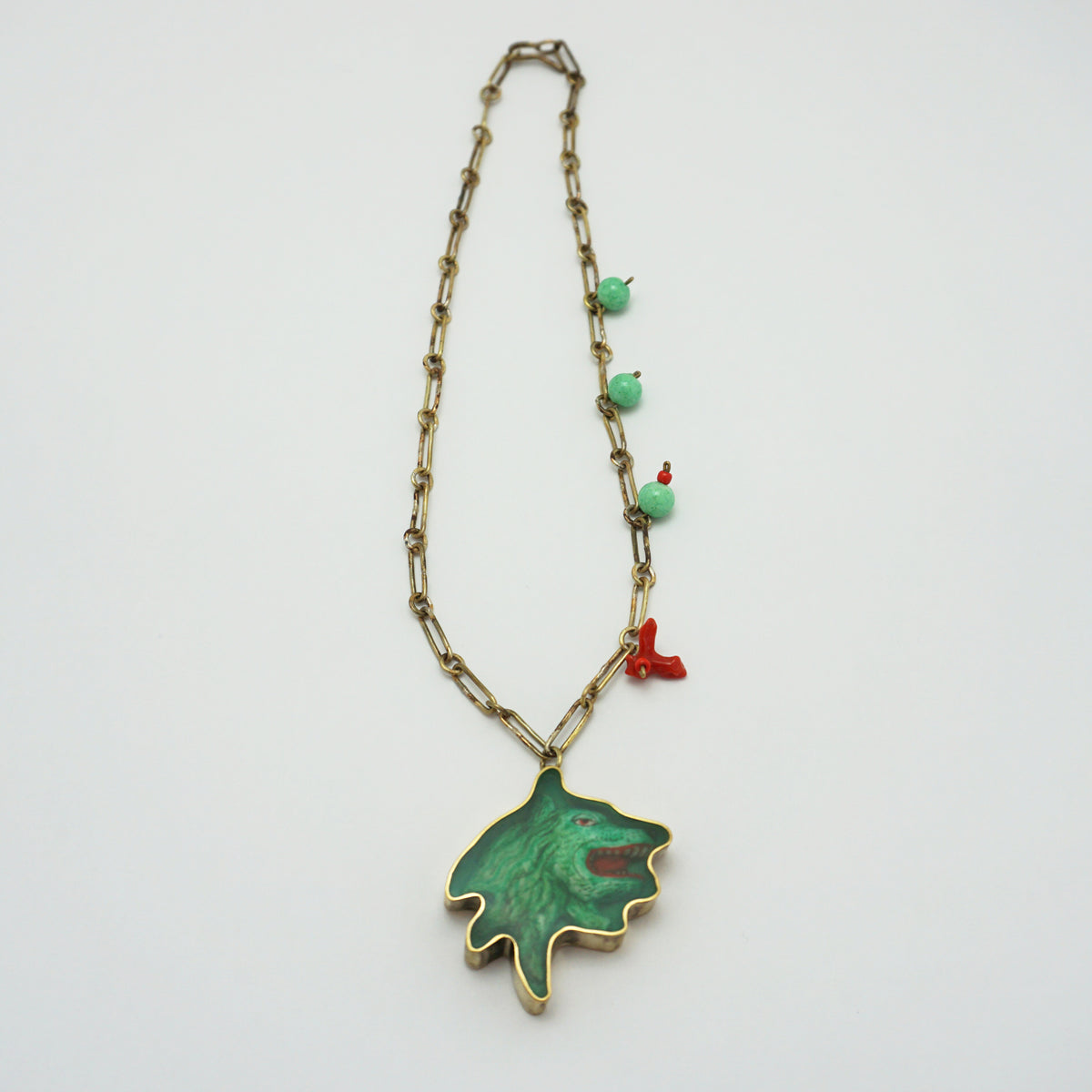 A vintage-style necklace featuring a gold-tone chain, small turquoise-green beads, a tiny red coral charm, and a large green pendant with a gold-tone outline, displayed on a clean white background.