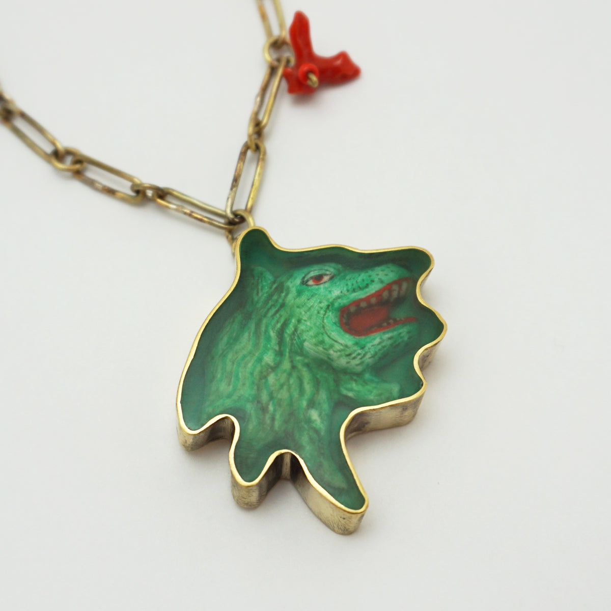 A gold-tone chain necklace with a pendant shaped like a green monster or creature head, featuring a textured green face, red mouth interior, and a small red coral charm on the chain, shown on a plain white background.
