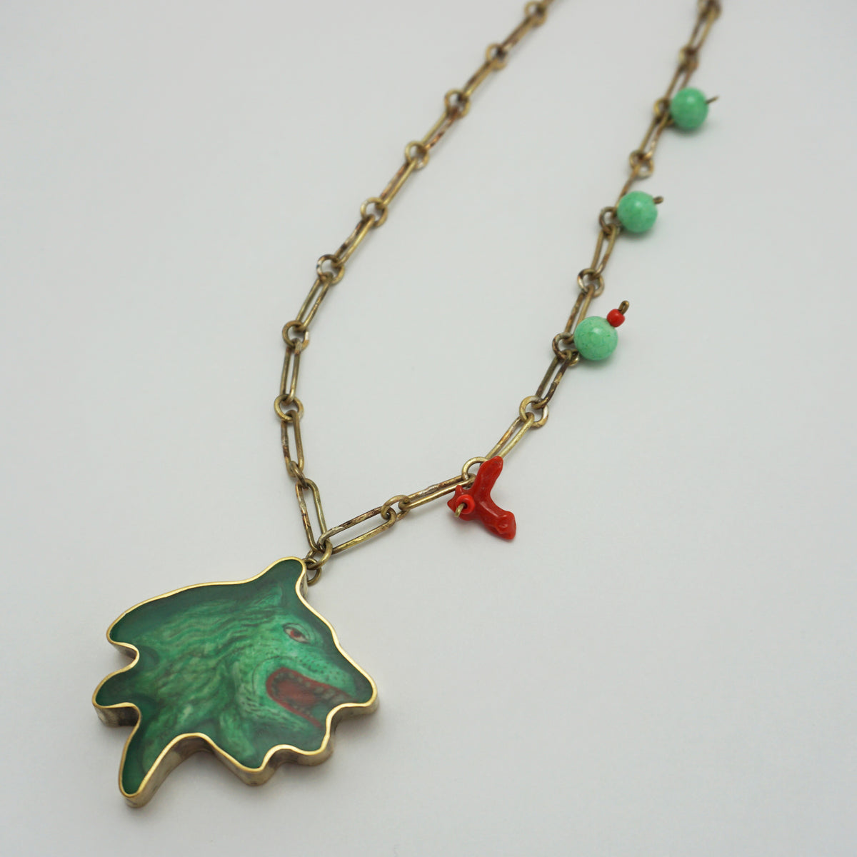 An antique-style necklace featuring a green pendant with gold trim, a small red coral charm, and green beads on an aged gold chain, shown on a plain light background.