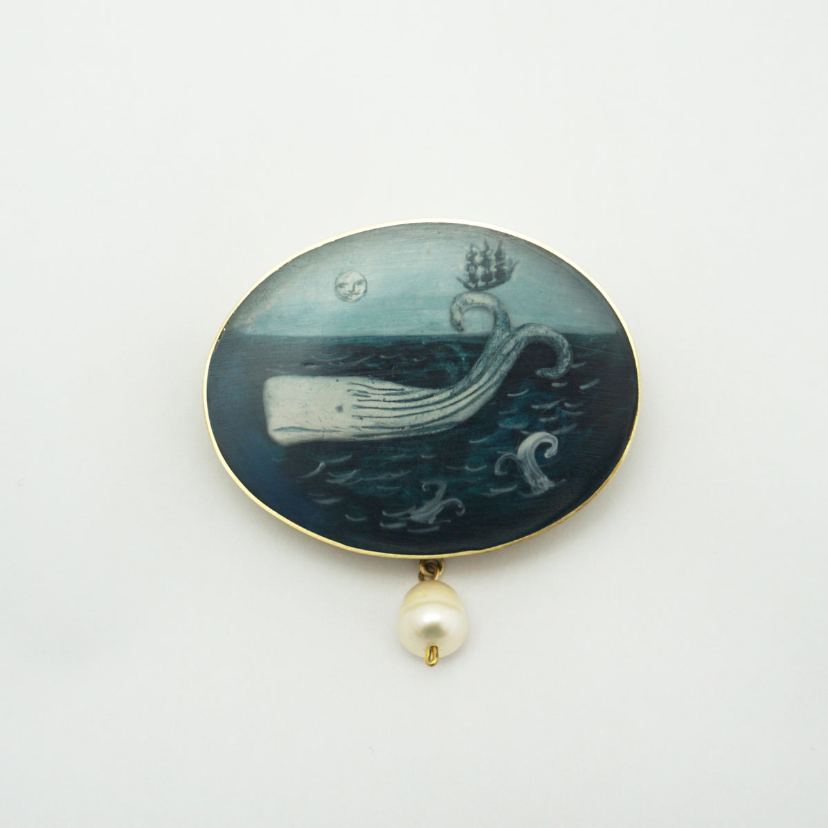 An oval brooch featuring a detailed hand-painted scene of a whale on the ocean waves under a dark night sky, with a single white pearl hanging below on a gold-tone chain.