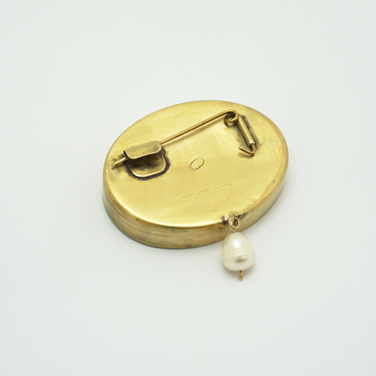 A gold oval pinback brooch showing the back side, featuring a small dangling pearl bead, set against a plain white background.