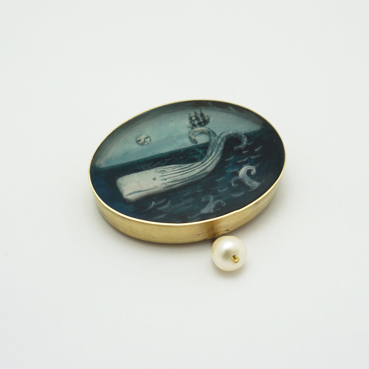 An oval gold-toned brooch featuring a painted whale scene in dark blue and grey tones, with a small white pearl bead hanging below it, photographed on a plain light background.