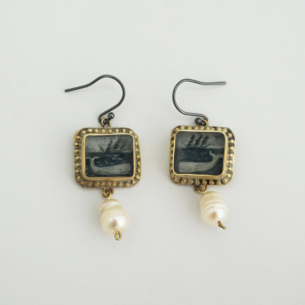 A pair of vintage-style drop earrings featuring square gold-toned frames with dark navy blue ship medallions, hanging from oxidised posts and finished with white pearl beads below, set against a plain light grey background.