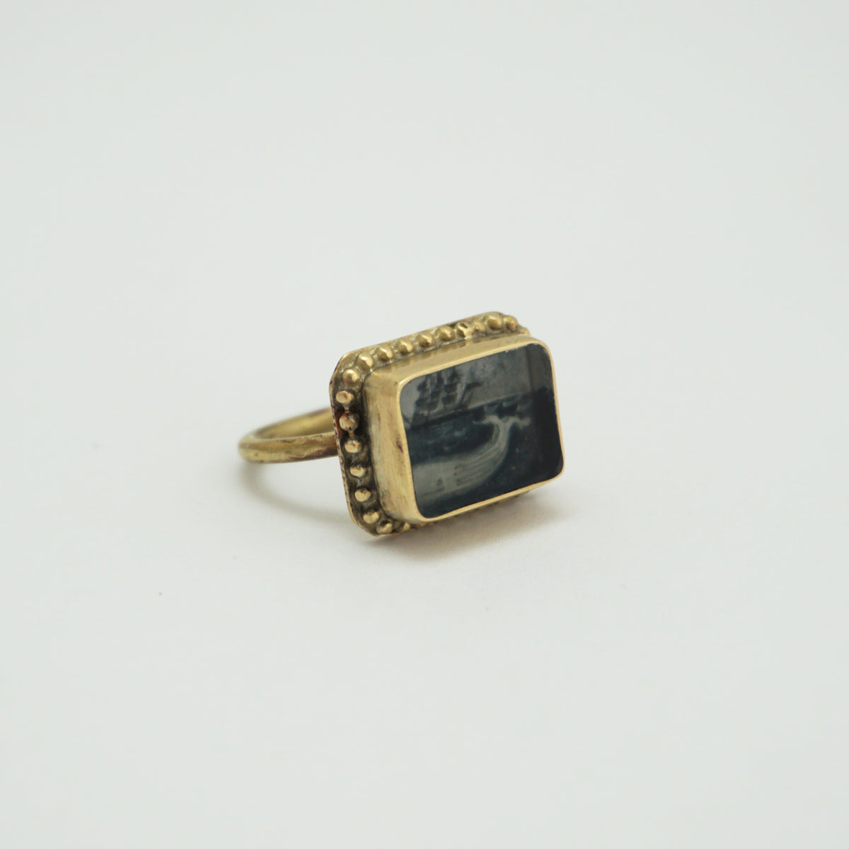 A side view of a gold rectangular ring with a hand painted whale, sea and ships. The outer part of the ring has circular bobbles around the edge.